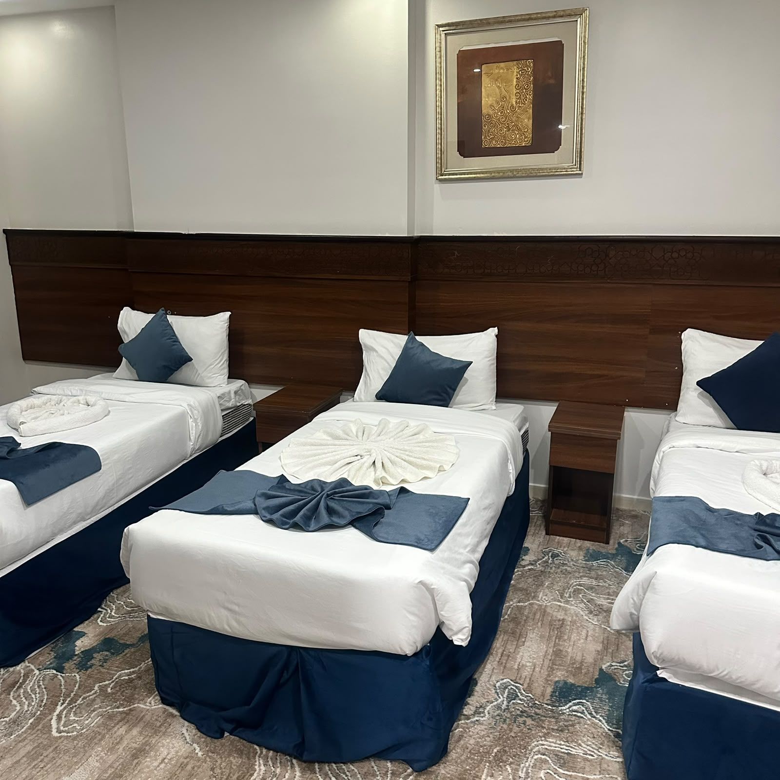 Een hotelkamer met drie bedden, elk met witte beddengoed en blauwe accenten, een houten hoofdeinde en een kunstwerk in een lijst aan de muur.