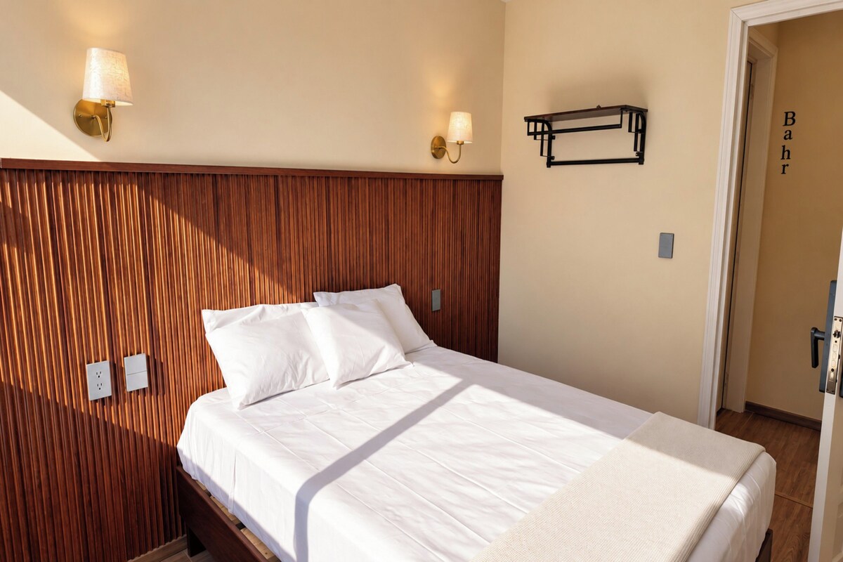 Una habitación de hotel con cabecera de madera, cama con ropa de cama blanca, dos lámparas fijadas a la pared y una puerta con pomo.