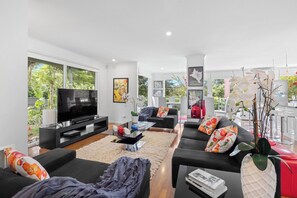 Ein modernes Wohnzimmer mit Flachbildfernseher, bequemen Sitzgelegenheiten und großen Fenstern mit Blick auf Grünflächen.