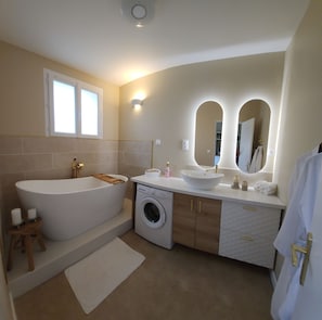 A modern bathroom with a freestanding bathtub, a built-in washing machine, and a vanity with a sink and mirror. - New Listing (Hure)