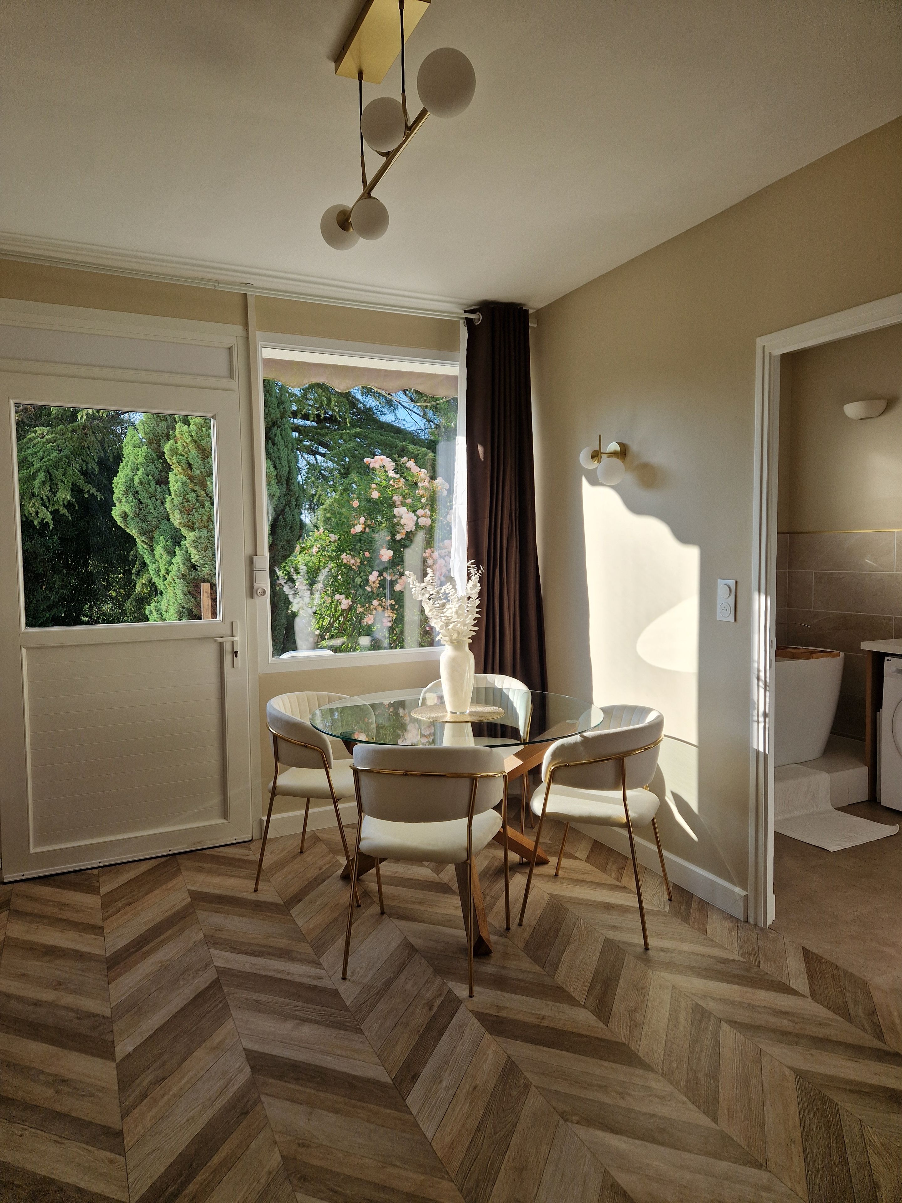 A dining area with a glass table, four chairs, a vase, and a view of greenery outside.