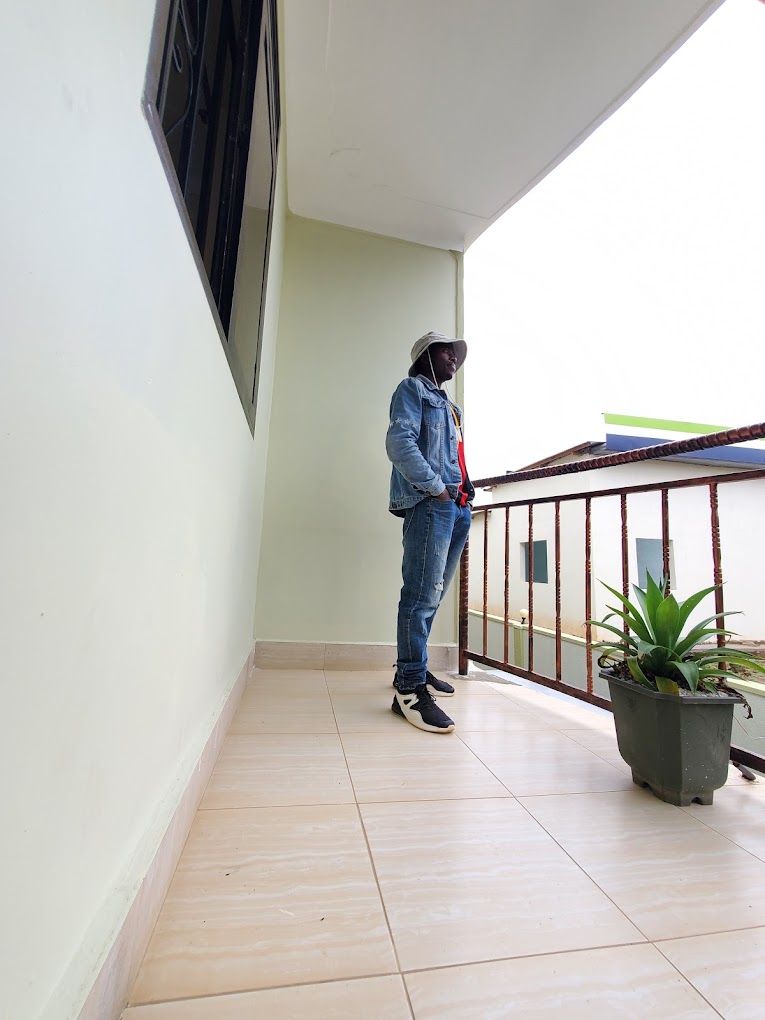 Une personne se tient sur un balcon, avec un pot de fleurs, une balustrade et une fenêtre.