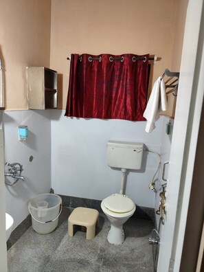 Une petite salle de bain comprenant des toilettes, un seau, un tabouret, une douche et un porte-serviettes.
