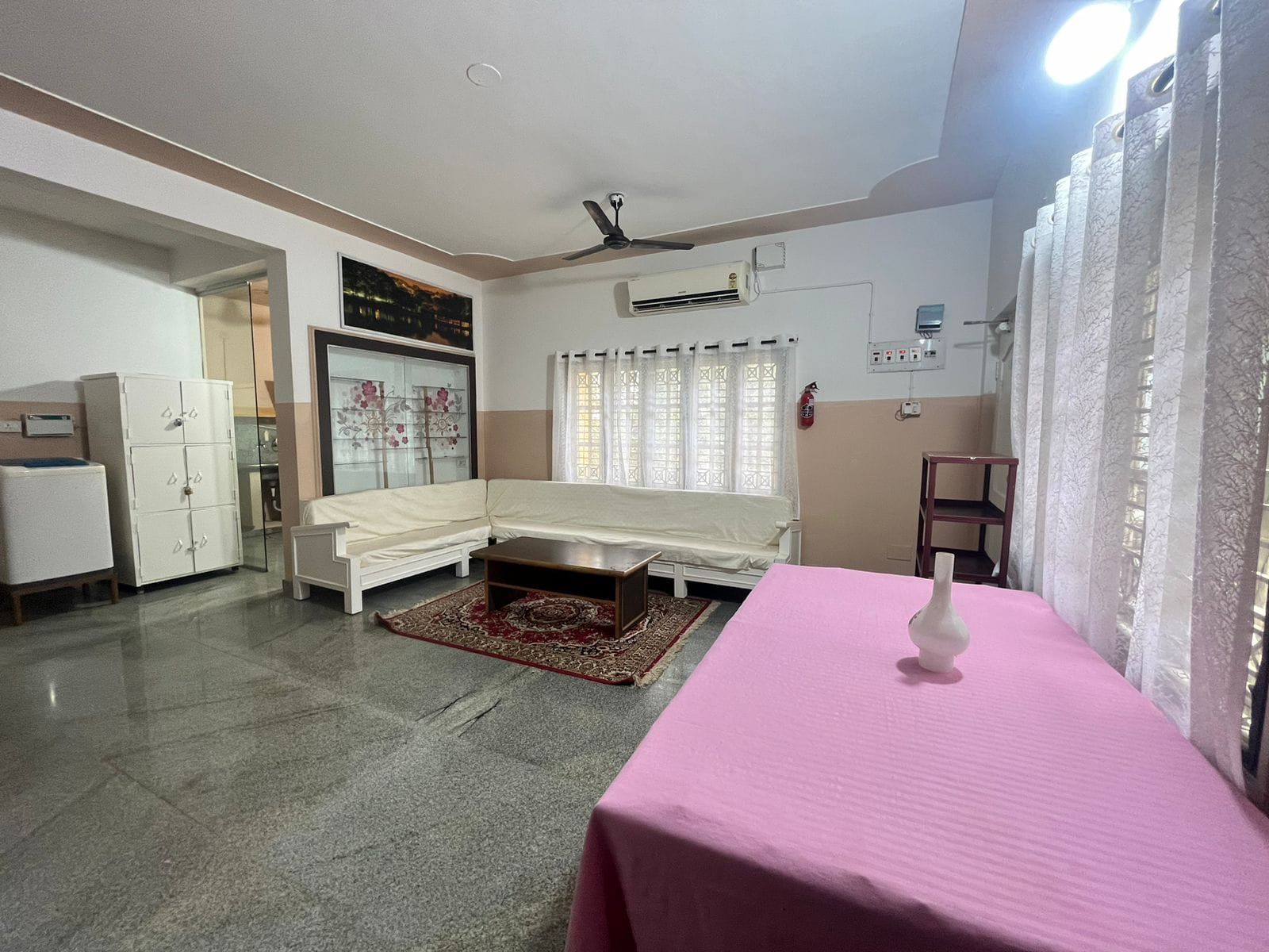 Una habitación con un sofá blanco, una mesa rosa, un ventilador de techo y una ventana con cortinas.