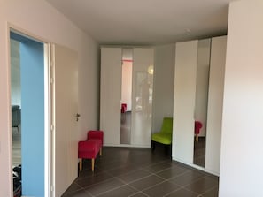 Un pasillo con una puerta que da a una habitación, una silla roja, una silla verde y un espejo que refleja a una persona.
