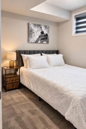 A bedroom with a bed, a nightstand, a lamp, a wall-mounted picture of the Eiffel Tower, and a window with blinds. - 
Airport luxury 1 bed suite (Calgary)