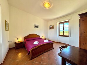 Une chambre à coucher avec un lit, des tables de chevet, une chaise en bois, une fenêtre donnant sur un paysage agréable et une porte.
