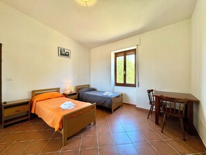 Une chambre avec deux lits simples, un bureau en bois, une chaise, une fenêtre donnant sur un beau paysage et une petite table avec une lampe.