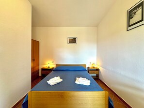 Une chambre d’hôtel étroite, avec un lit simple, deux tables de chevet avec des lampes, une armoire en bois et un tableau encadré au mur.