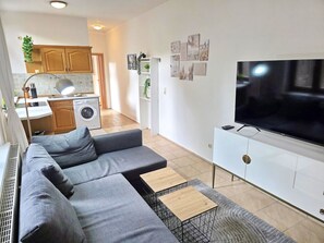 Ein modernes Wohnzimmer mit einem grauen Ecksofa, einem Flachbildfernseher auf einem weißen Schrank und einer Küche im Hintergrund.