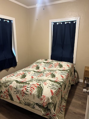 Une chambre à coucher avec un lit, deux fenêtres avec des rideaux, une chaise en bois et une porte.