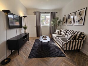 A modern living room with a black sofa, a coffee table, a TV on the wall, and a window with curtains. - 3 bedroom home in Horsham (Southwater)