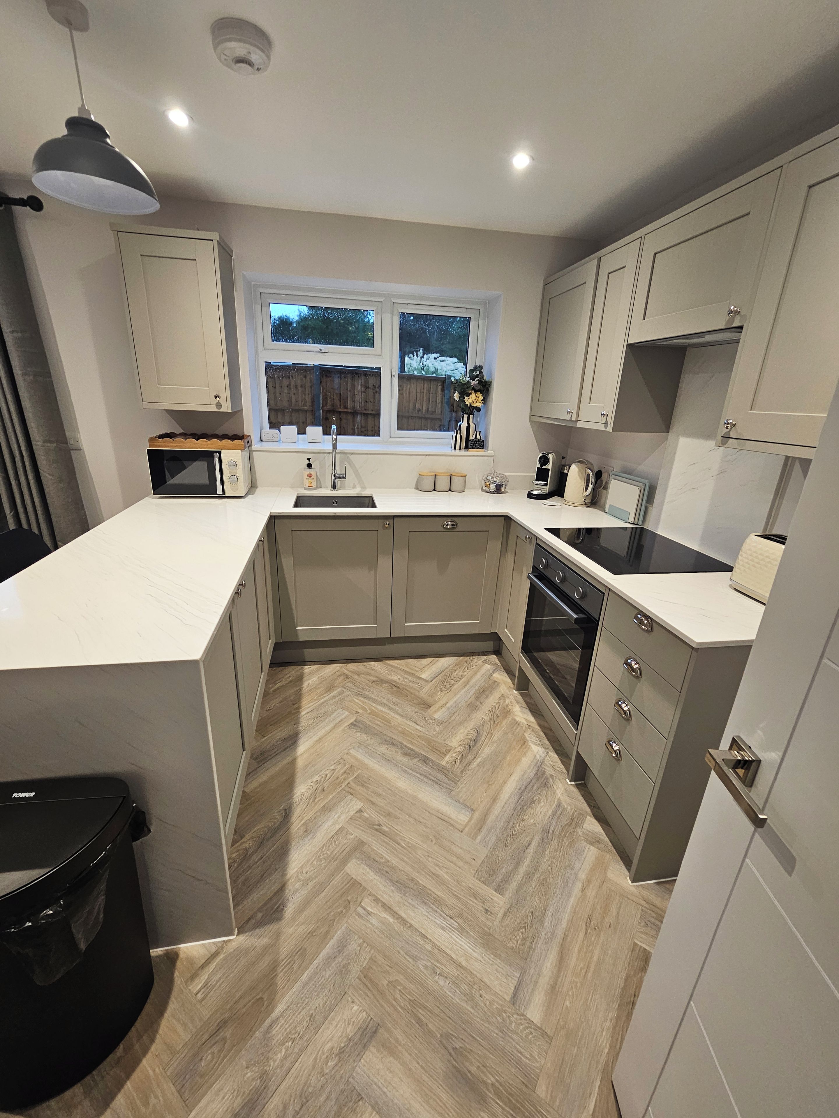 Une cuisine moderne avec un parquet en bois disposé en chevrons, des meubles blancs, un four encastré et une fenêtre donnant sur l’extérieur.