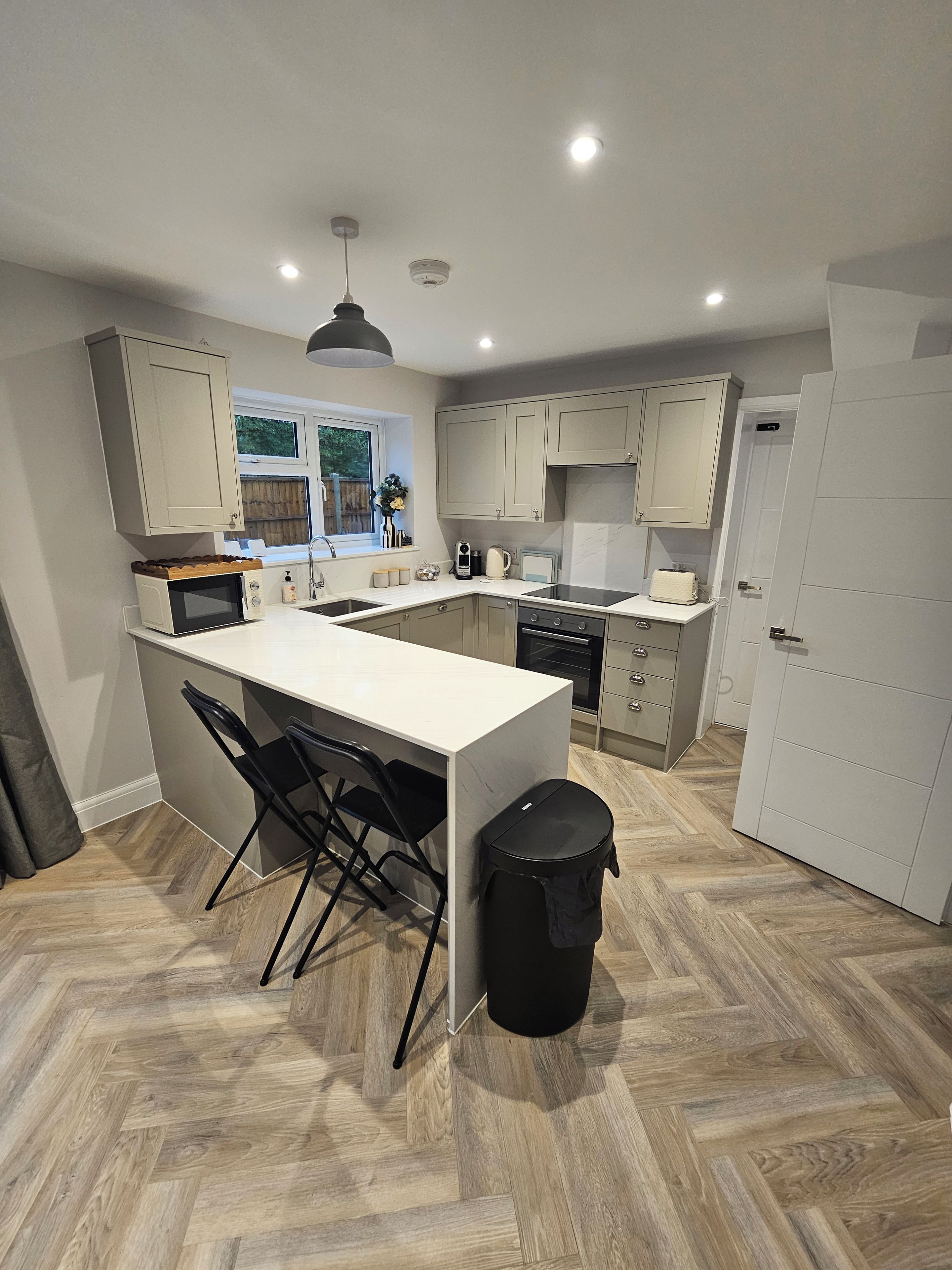 A modern kitchen with a central island, black chairs, a trash bin, and a window with a view.