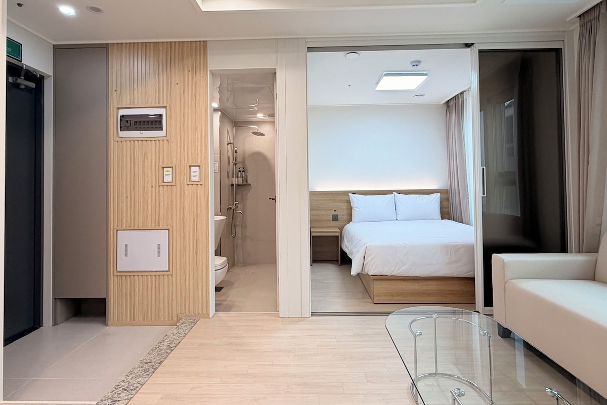 Moderni hotellihuone, jossa on sänky, lasinen sohvapöytä, sohva, suihkutila ja ovi, joka johtaa toiseen huoneeseen.
