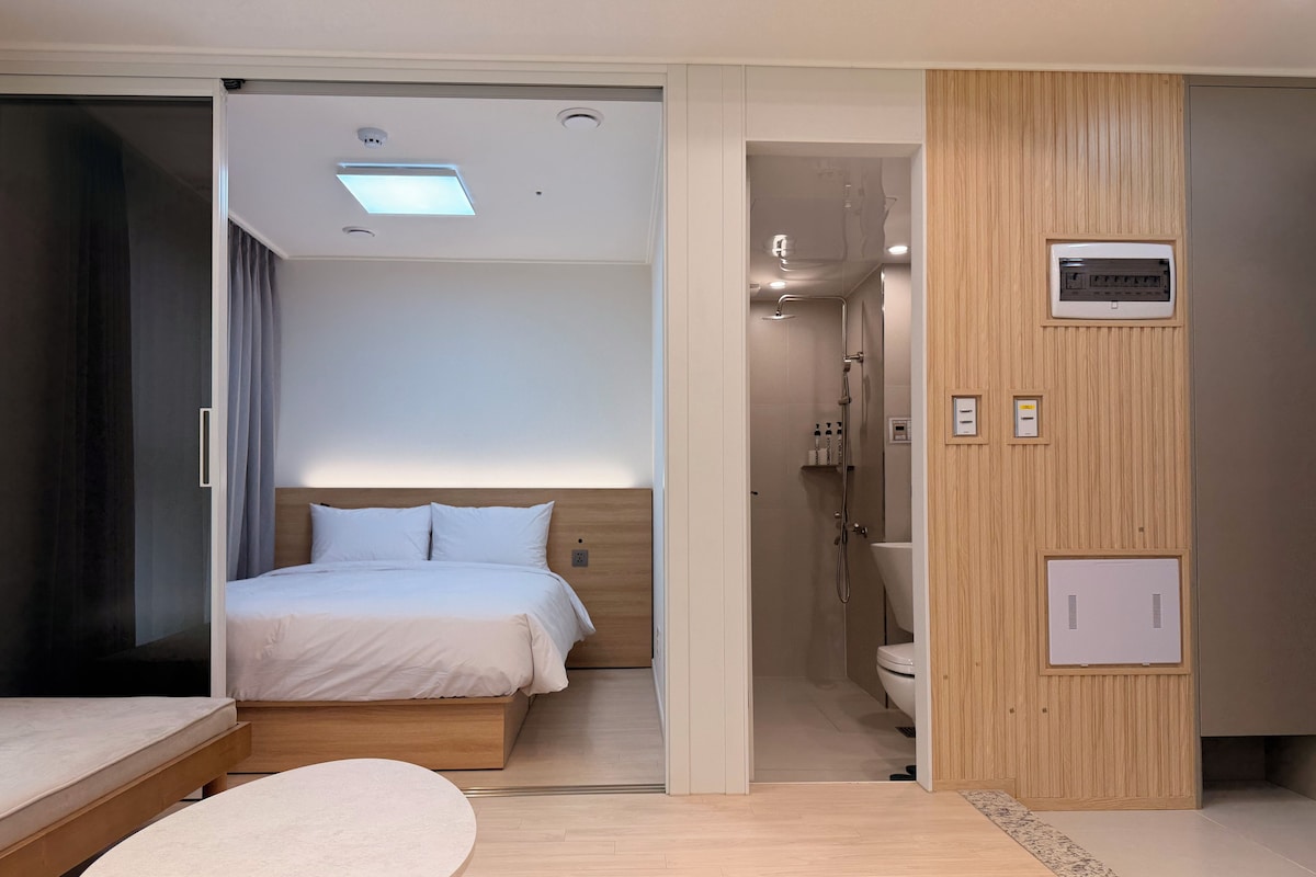 Moderni hotellihuone, jossa on sänky, pieni pöytä, suihkutila ja puupanelilla verhoiltu seinä.