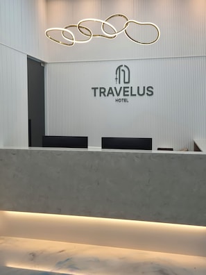 Modernilla designilla varustettu vastaanottotiski, jossa on hotellin, TRAVELUS HOTEL, logo ja ainutlaatuinen kattovalaisin.