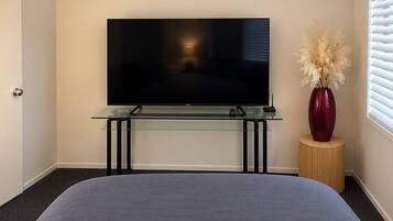 A bedroom with a bed, a TV on a glass stand, a vase with dried plants, and a window with blinds.