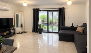 A living room with a flat-screen TV, a sofa, a coffee table, and a sliding glass door leading to a balcony with plants. - Cosy suburban townhouse (Acacia Ridge)