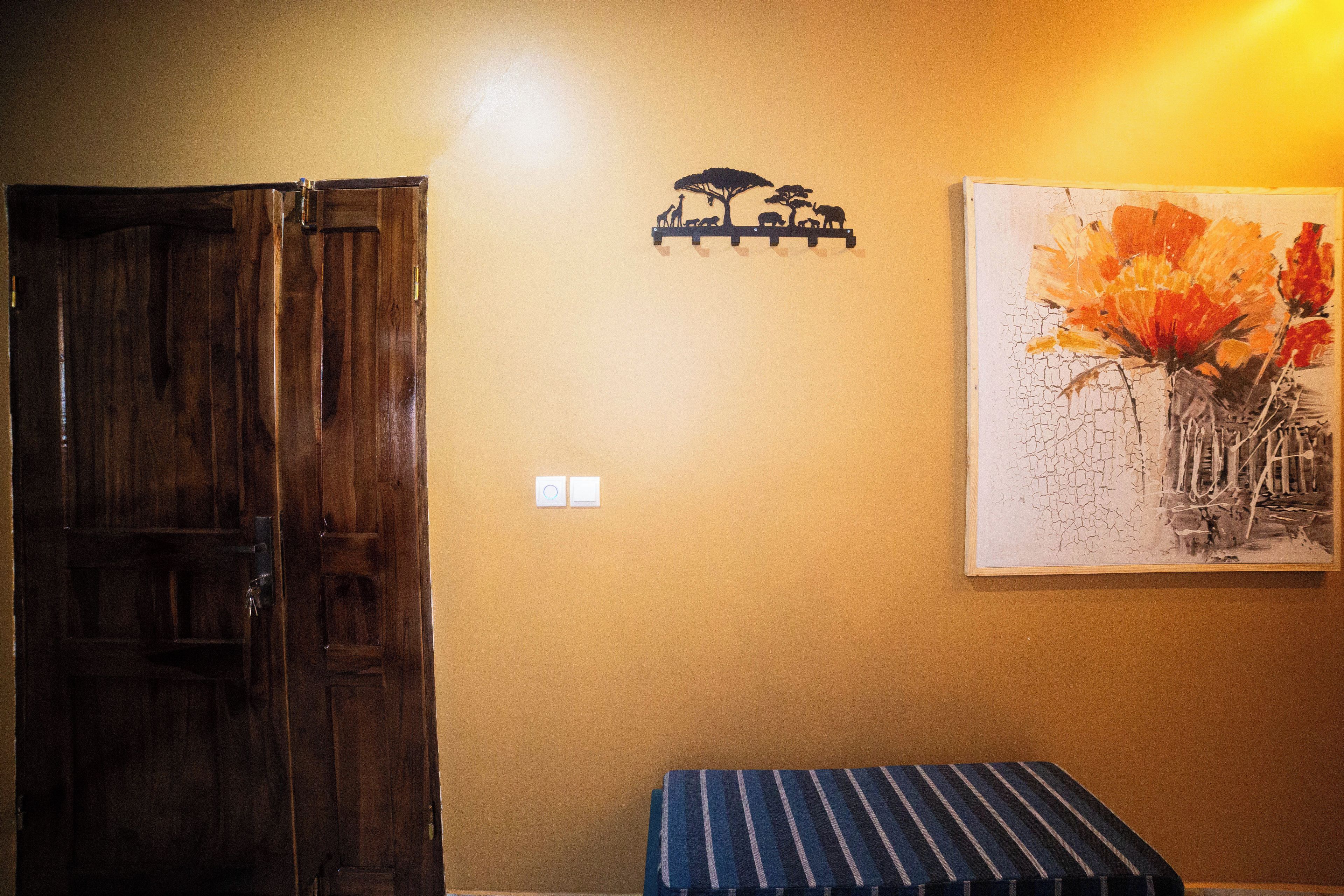 A room with a wooden door, a wall-mounted light switch, a small shelf with a silhouette decoration, and a framed painting of flowers.