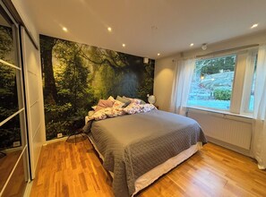 Quarto com uma cama grande, um mural com tema de floresta na parede e uma janela com cortinas.