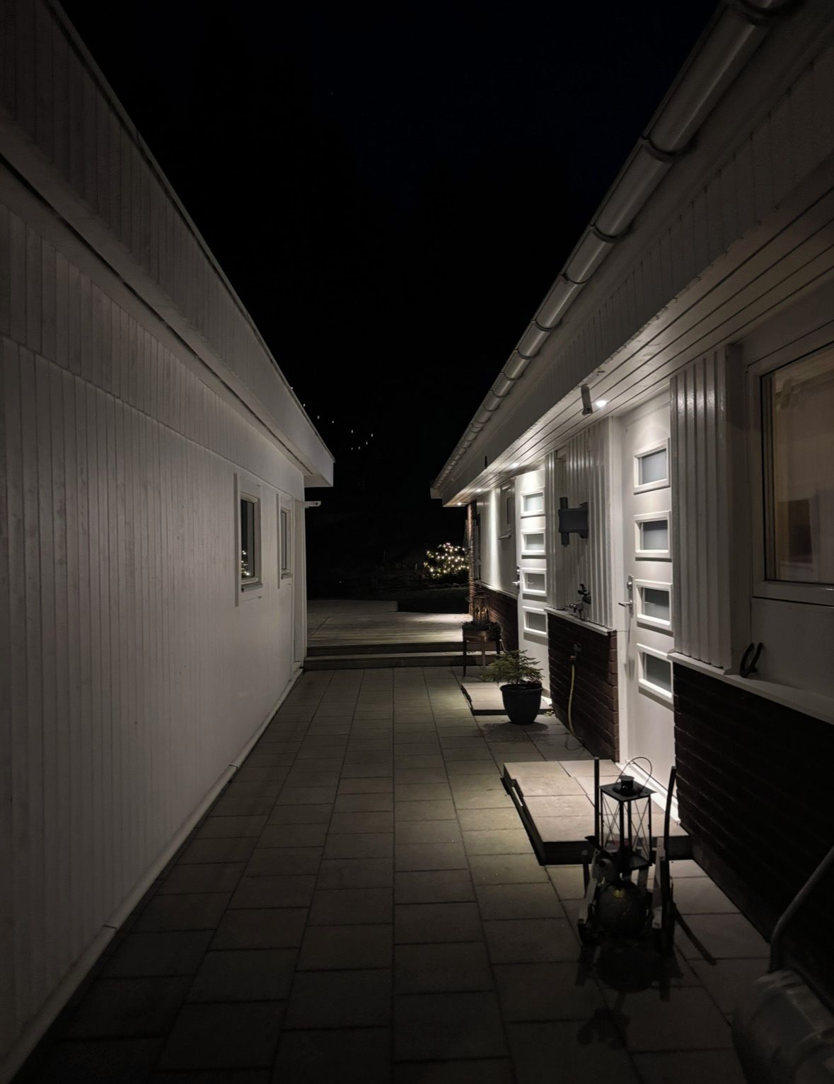 Un pasillo estrecho con piso de baldosas, bordeado por edificios blancos con ventanas y puertas, que conduce a una zona más luminosa con un banco y una planta en maceta.