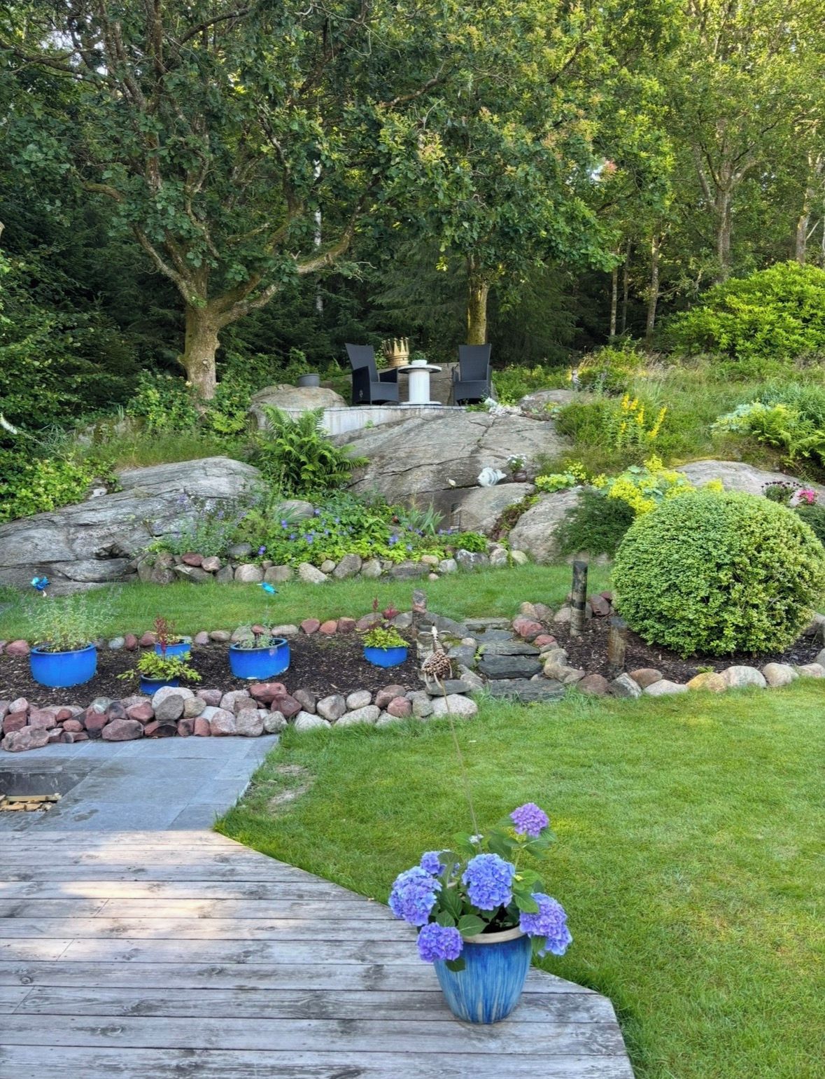Um jardim com deck de madeira, vasos de plantas e área de estar com cadeiras e mesa.