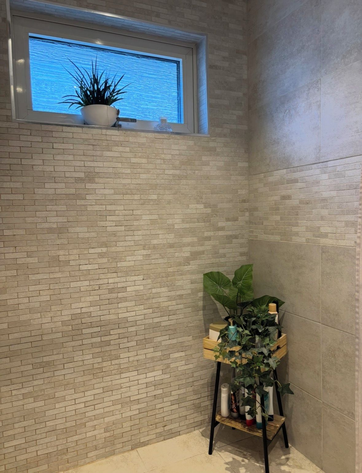 Una habitación de esquina con pared de ladrillo, una ventana pequeña con una planta y un estante de madera con planta en maceta.