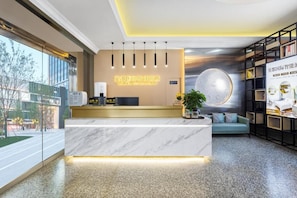 Zona de recepción de hotel con mostrador de mármol, un escritorio, una gran decoración circular en la pared y un área de descanso con sofá y planta.