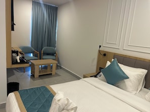 Habitación de hotel con una cama, un escritorio, dos sillas, una mesita, un cesto de basura y una ventana con cortinas.