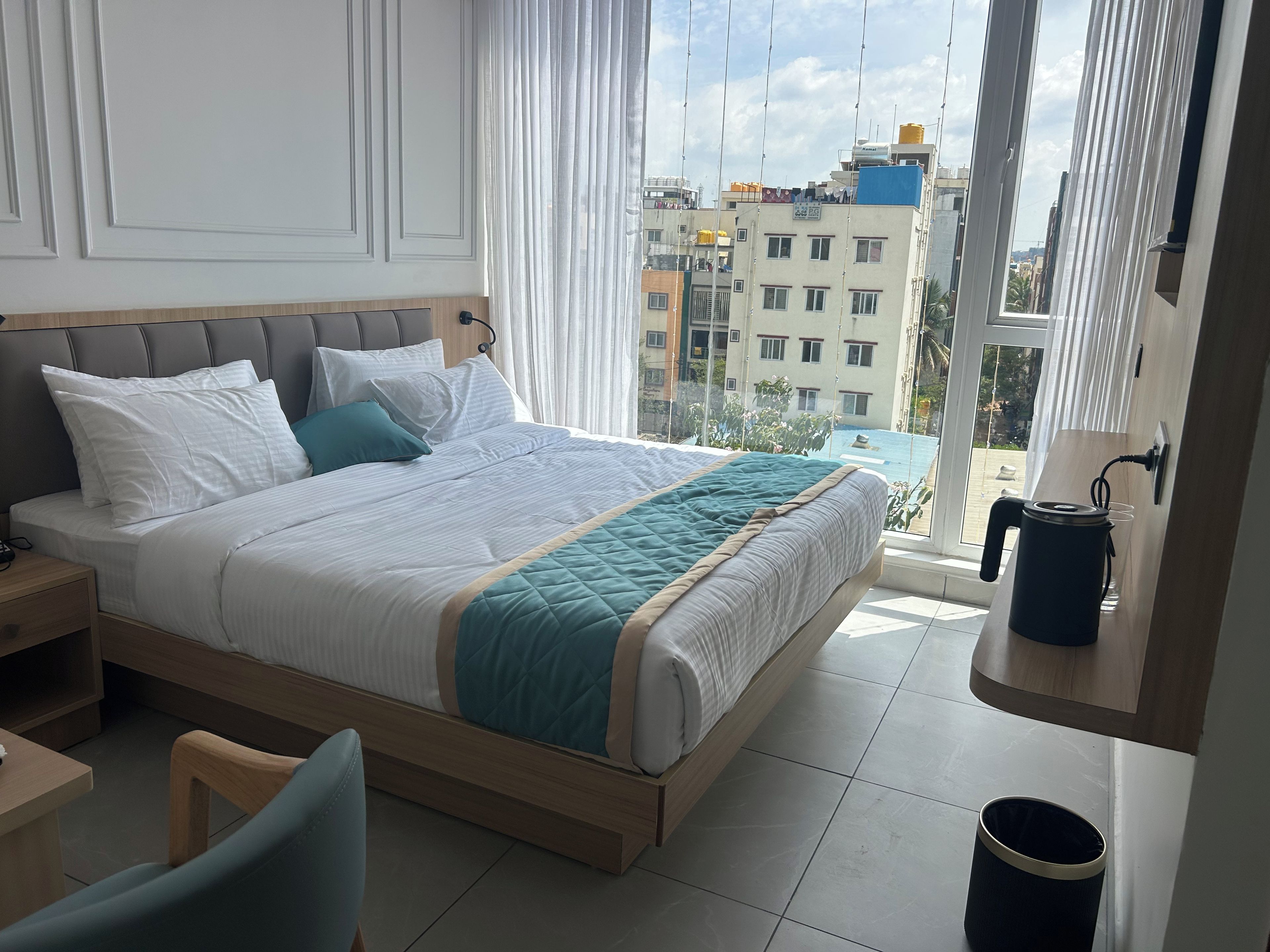 Ein modernes Hotelzimmer mit einem großen Bett, einem Nachttisch, einem Sessel und Blick auf die Stadt durch große Fenster.