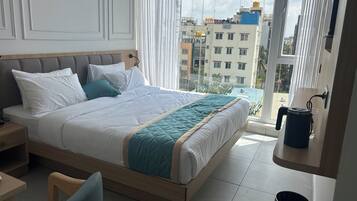 Ein modernes Hotelzimmer mit einem großen Bett, einem Nachttisch, einem Sessel und Blick auf die Stadt durch große Fenster.