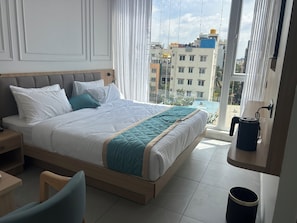 Quarto de hotel moderno com uma cama grande, mesa de cabeceira, uma cadeira e vista para o horizonte urbano pelas amplas janelas.