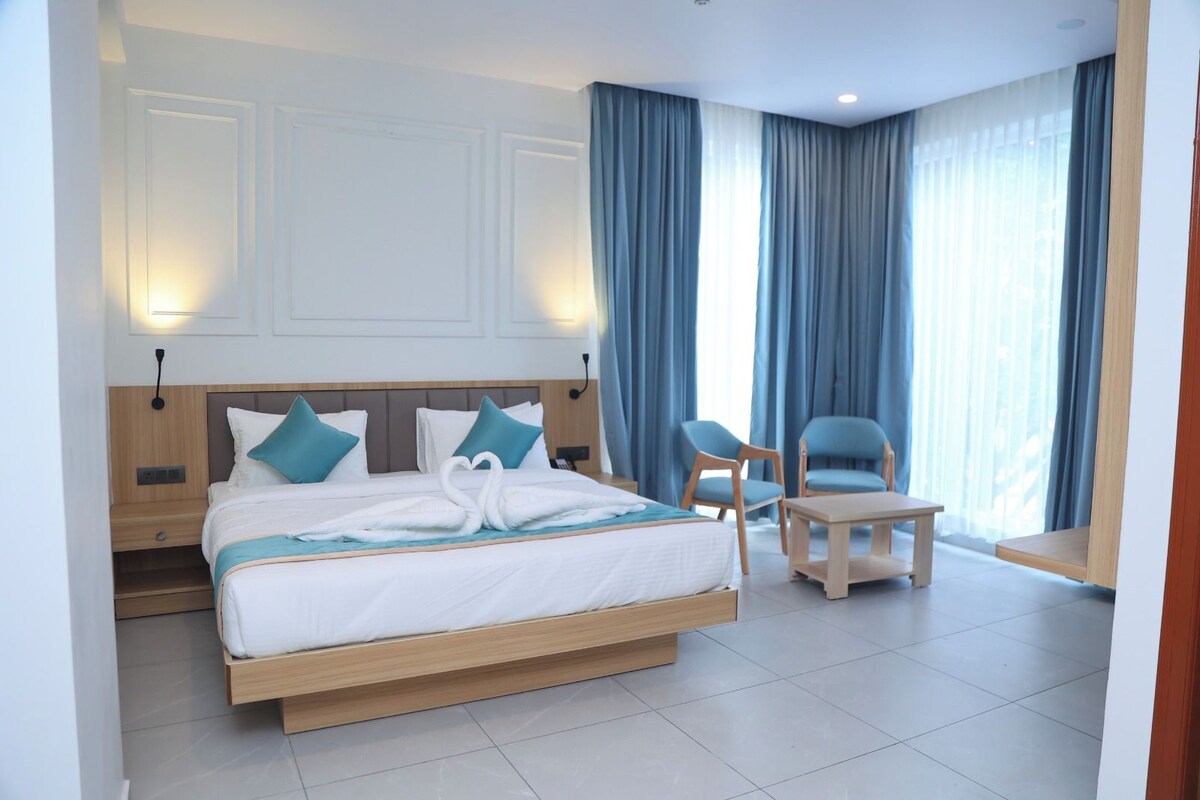 Quarto de hotel com uma cama grande, duas cadeiras, uma mesinha e vista para o exterior através de cortinas.