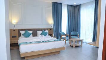 Ein Hotelzimmer mit einem großen Bett, zwei Stühlen, einem kleinen Tisch und Blick ins Freie durch Vorhänge.