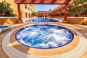 Un bain à remous entouré de chaises longues et une piscine, avec un bâtiment en arrière-plan.
