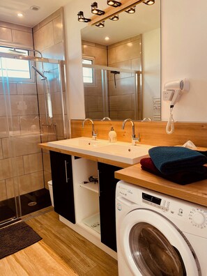 Ein modernes Badezimmer mit Duschkabine aus Glas, einem Waschbecken mit Holzbeschlag, einem Spiegel, einem Fön und einer Waschmaschine.