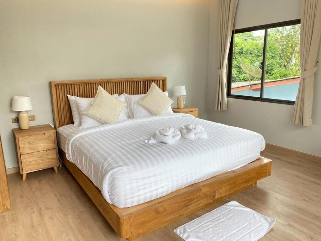 A bedroom with a wooden bed frame, white bedding, two bedside tables with lamps, and a window with a view of greenery.