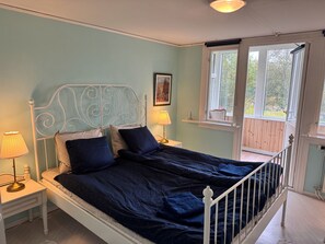 Ein Bett mit Metallkopfteil, zwei Nachttische mit Lampen, ein Fenster mit Blick ins Grüne und eine Tür zu einem Außenbereich.