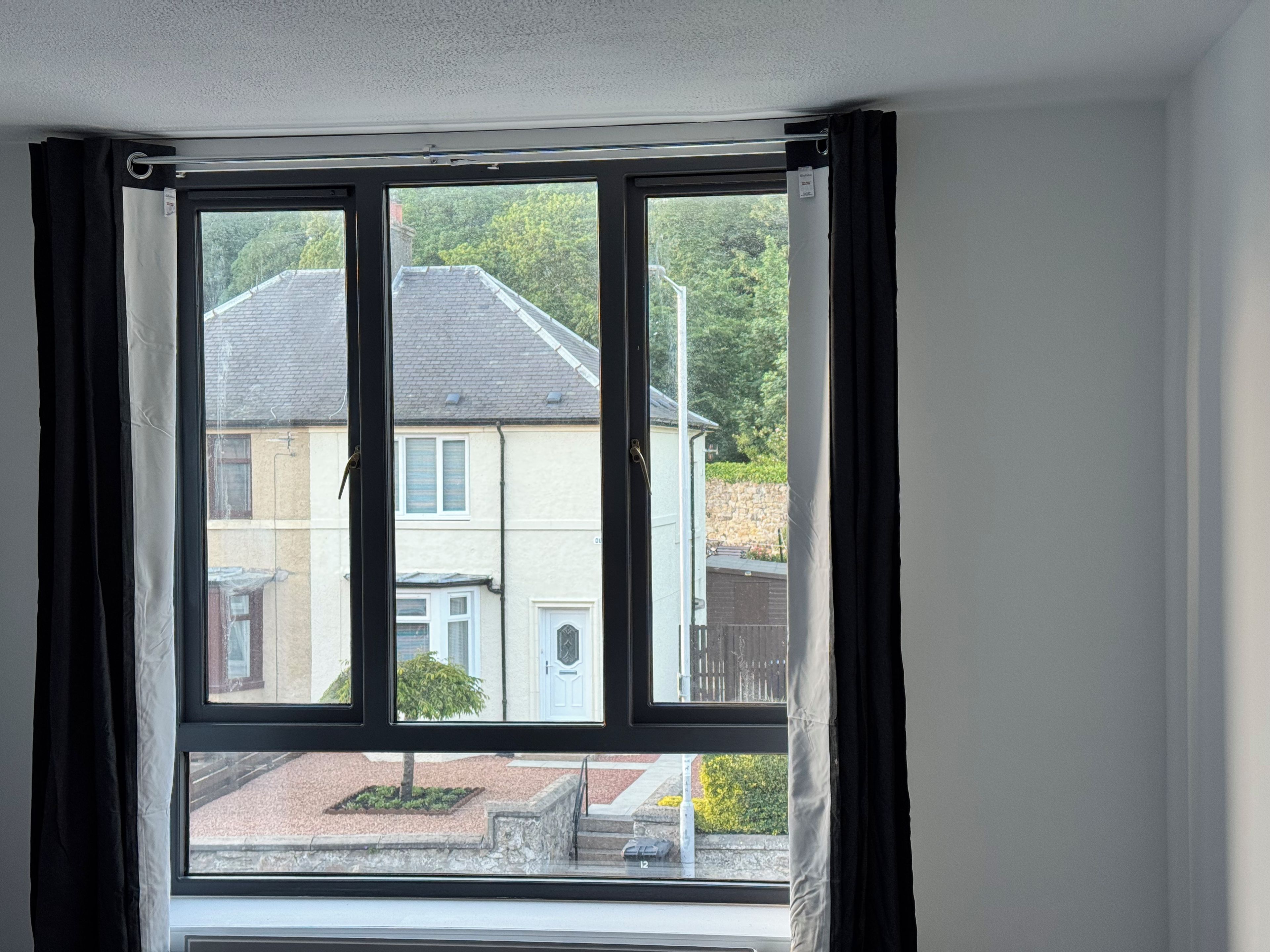 A room with a large window, a radiator, and a view of a house and greenery outside.