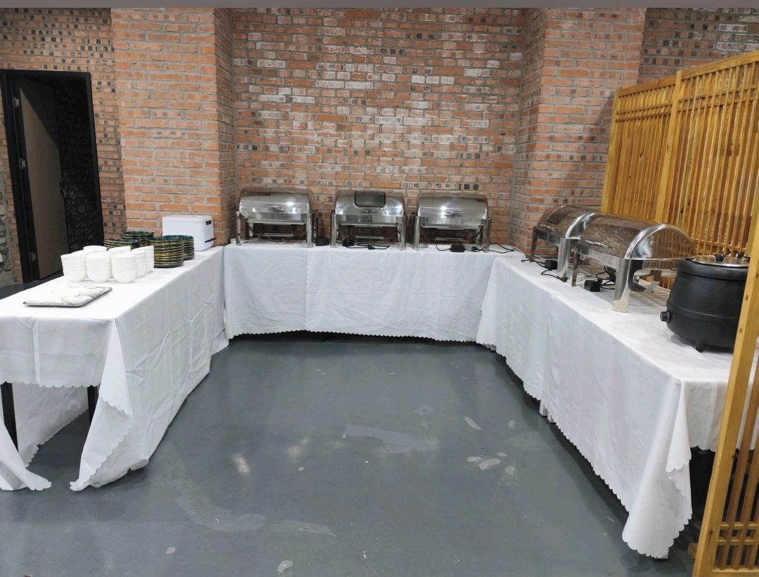 Montaje de bufé con múltiples dispensadores de comida, una mesa cubierta con mantel blanco y un fondo de pared de ladrillo.