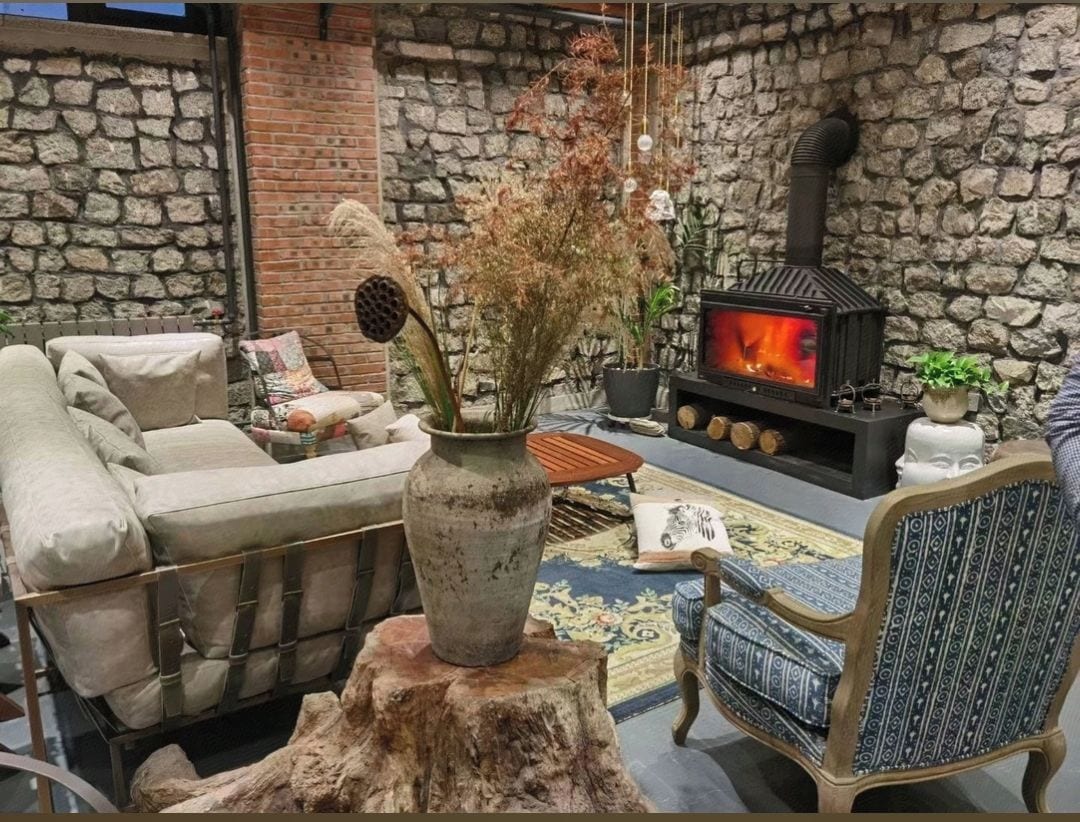 Ein gemütlicher Wohnbereich mit einer Steinwand, einem Sofa, einem gemusterten Sessel, einer Vase mit Trockenpflanzen und einem Kaminofen.