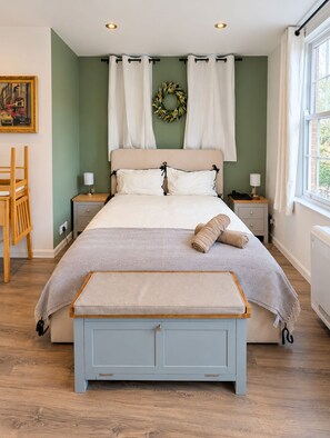 A bedroom with a bed, two bedside tables, a bench, a chair, a wall-mounted wreath, and a window with curtains.