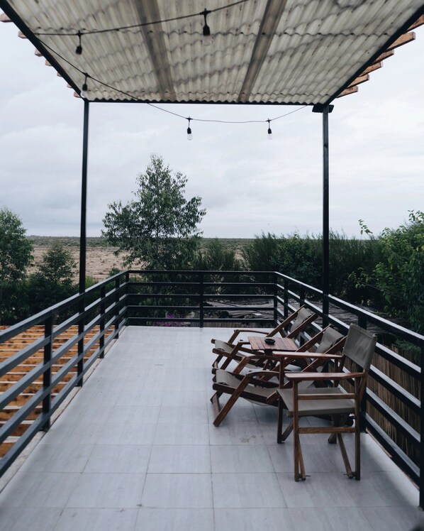 A covered outdoor seating area with wooden chairs and tables, a metal railing, and a view of the landscape.