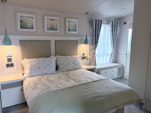 A neatly arranged bedroom with a bed, bedside tables, a window with curtains, and wall-mounted artwork. - Ambleside Lodge Sleeps 6 With Parking and Wrap Around Balcony (Porthmadog)