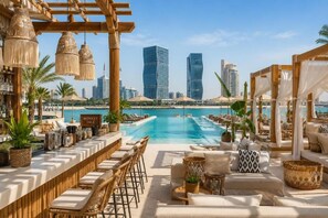 A poolside bar area with lounge chairs, a bar counter, and a view of modern skyscrapers in the background. - Zigzag Tower B-Travellers' Retreat, 2 BR with Pool and Sea View at Pearl-1008 (Lusail)