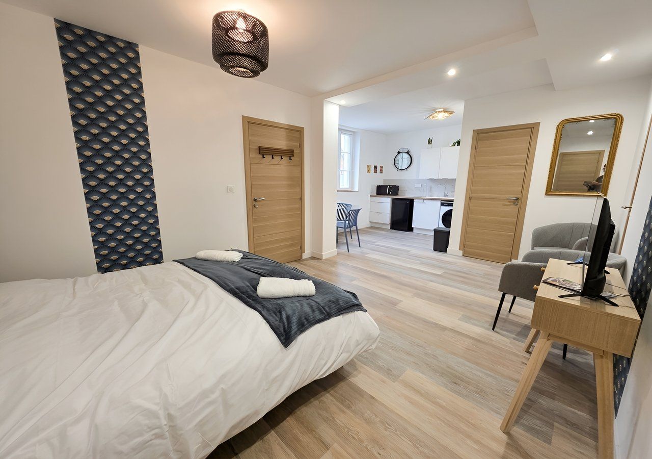 Ein Hotelzimmer mit einem Bett, einem Schreibtisch, einem Sessel, einem Spiegel und einer Küchenzeile im Hintergrund.