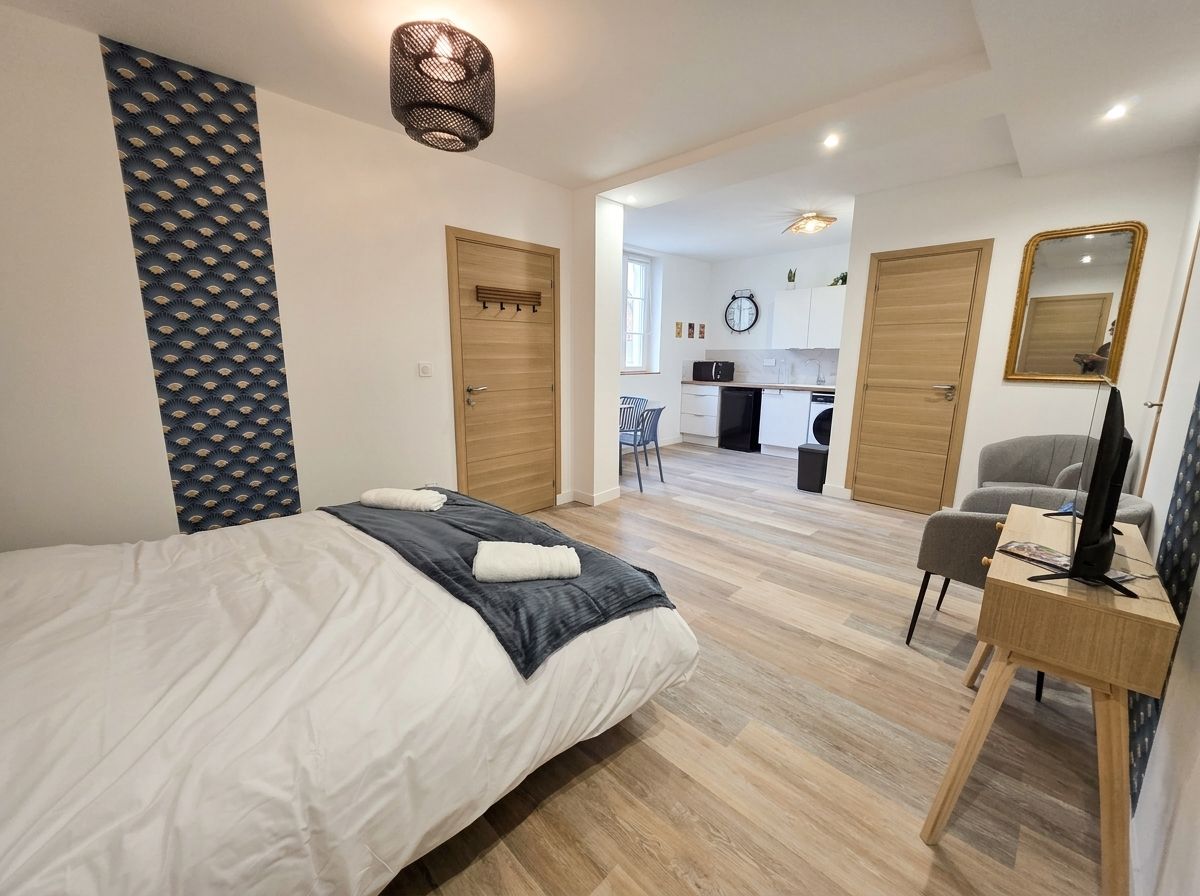 A bedroom with a bed, a desk, and a chair. There is a kitchenette in the background.