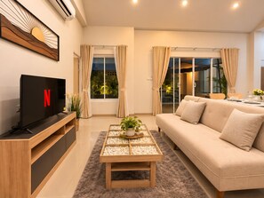 Een moderne woonkamer met een flatscreen televisie, een bank, een salontafel met decoratieve objecten en uitzicht op het groen door grote ramen.