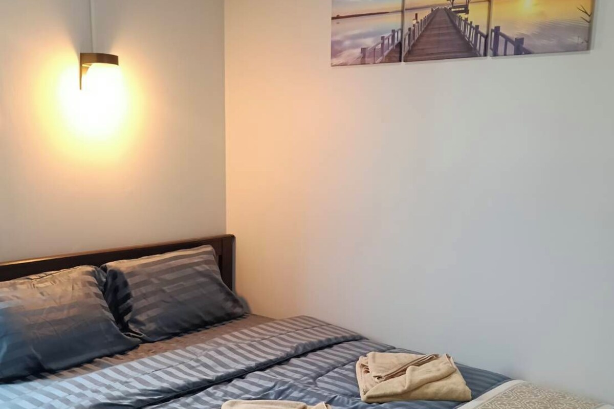 A bedroom with a bed, pillows, a blanket, a wall-mounted light, and a sunset painting.
